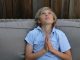 Boy looking to the sky, smiling face and folded hands (prayer will be joy). He is wearing a blue shirt and is sitting on a gray sofa.