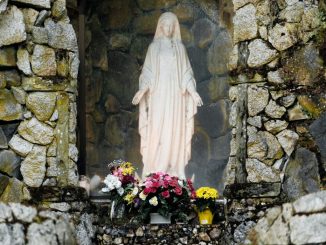 Statue of the Virgin Mary in an alcove of a gray stone wall. Yellow, red and white flowers at the foot of the statue.