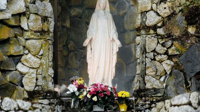 Statue of the Virgin Mary in an alcove of a gray stone wall. Yellow, red and white flowers at the foot of the statue.