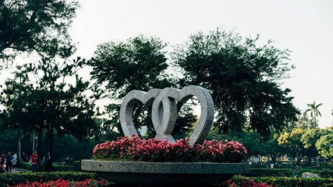 Fountain in a park, made of two intertwined concrete hearts (love without fear), surrounded by red flowers and greenery.