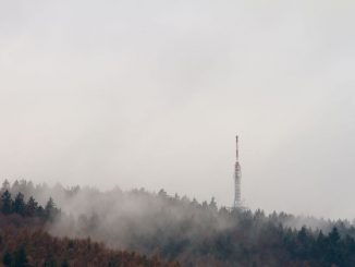 Transmission tower in a forest of green trees under white clouds during daytime.