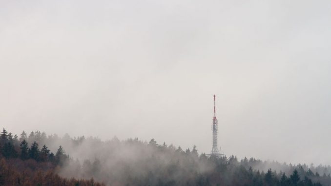Transmission tower in a forest of green trees under white clouds during daytime.
