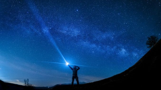 Silhouette of a man pointing a flashlight to the sky in the foreground. Starry blue sky in the background.