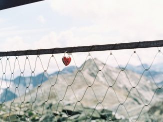 Heart shaped padlock suspended in a chain link fence. Mountains and white clouds in the background.