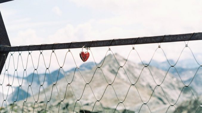 Heart shaped padlock suspended in a chain link fence. Mountains and white clouds in the background.