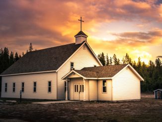 Small white and brown wooden church in the foreground. Coniferous forest in the background under partly cloudy sky during daytime.