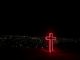 Red neon light cross in the foreground. Thousands of bright spots of a city under a completely black sky in the background.