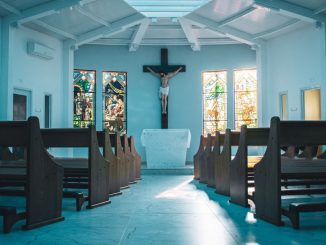 Dark brown wooden church pews in the foreground. Behind the small altar, a crucifix in the center and two stained glass windows on each side. Sky blue walls, ceiling and floor.