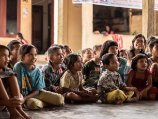 Group of children, sitting on ground, attentive and smiling, listening to the story of a person who cannot be seen.