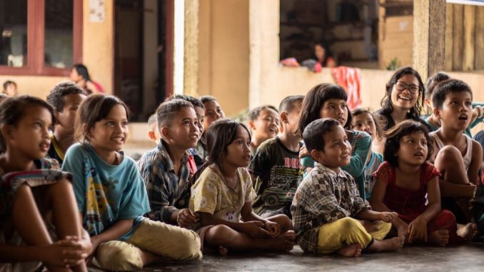 Group of children, sitting on ground, attentive and smiling, listening to the story of a person who cannot be seen.