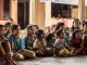 Group of children, sitting on ground, attentive and smiling, listening to the story of a person who cannot be seen.