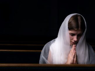 Women kneeling in a church pew, folded hands, eyes closed and white scarf on her head.