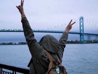Person wearing hoodie and backpack making peace signs with hands near body of water.