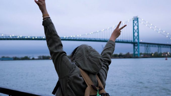 Person wearing hoodie and backpack making peace signs with hands near body of water.