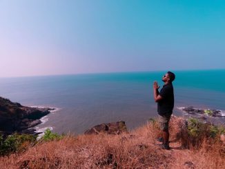 Man in black t-shirt, with folded hands, standing on brown rock formation near blue sea during daytime.