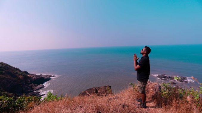 Man in black t-shirt, with folded hands, standing on brown rock formation near blue sea during daytime.