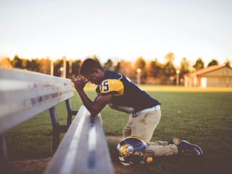 Baseball player kneeling on ground, folded hands and elbows on a bench during the day.