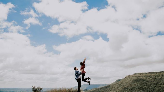 Man lifting woman on top of a mountain during daytime. Sky strewn with big white clouds.