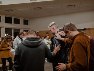 People in black and brown jackets standing in front of other people to pray in a hall.