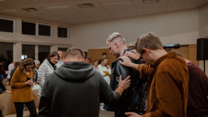 People in black and brown jackets standing in front of other people to pray in a hall.