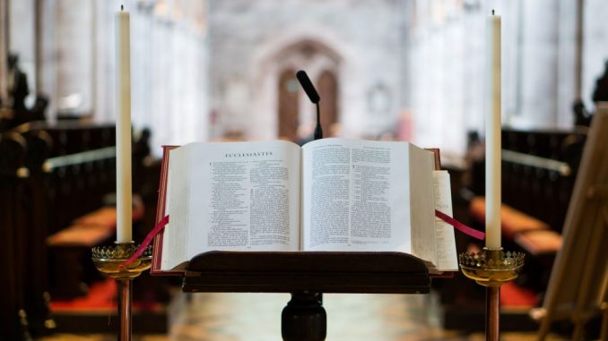 Open Bible on a dark brown wooden book stand and a long candle on each side.