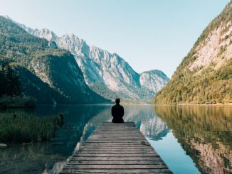 Man sitting on a gray dock facing a lake surrounded by high mountains.