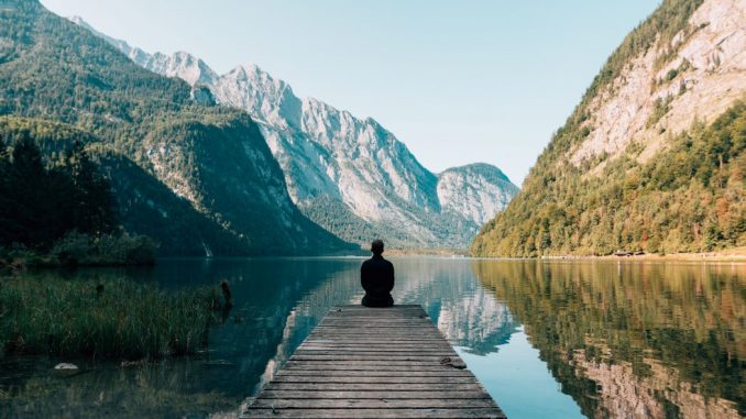 Man sitting on a gray dock facing a lake surrounded by high mountains.