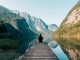 Man sitting on a gray dock facing a lake surrounded by high mountains.