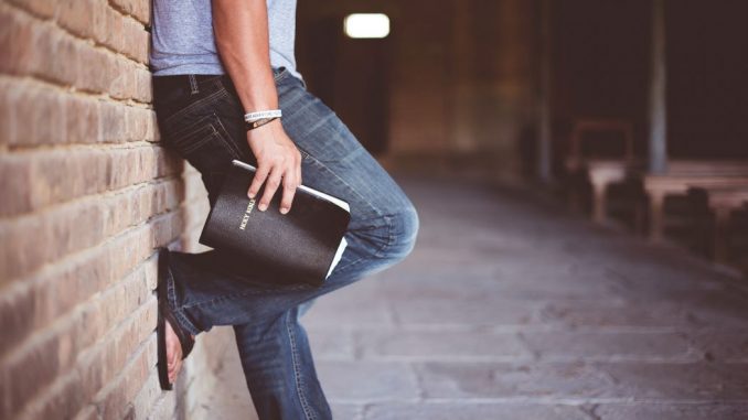 Man holding Holy Bible leaning on bricked wall.
